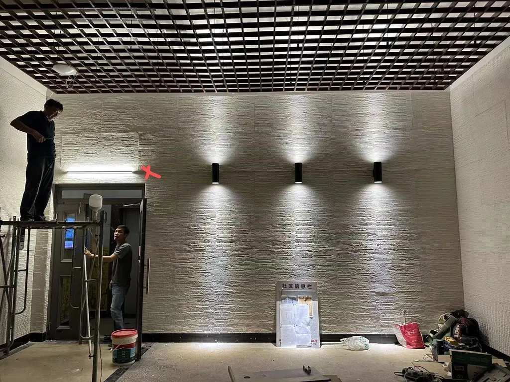施工中的社區大廳，三盞壁燈已裝上並點亮，在石材牆面上洗出三道光弧，工班仍在現場